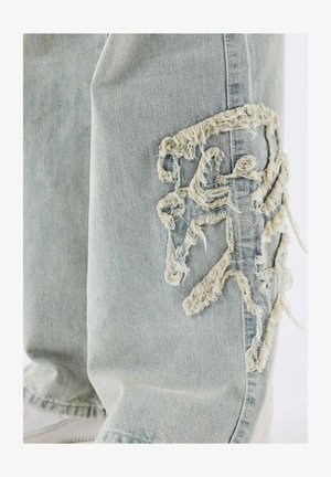 Jeans de mezclilla azul claro con bordes deshilachados y un diseño bordado que asemeja una figura en la parte izquierda de la rodilla.