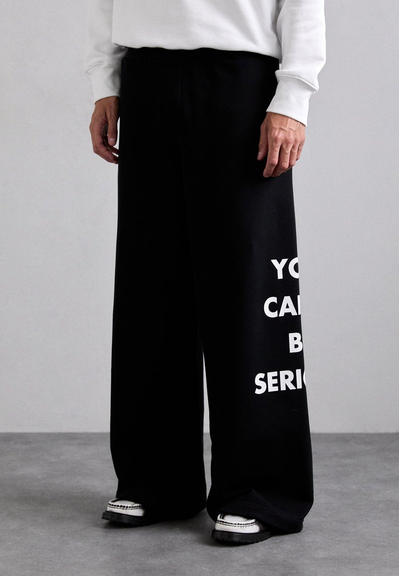 Čierne široké teplákové nohavice s jemnou textúrou, na ľavej nohe so sprievodným tučným bielym textom "NEPREHÁŇAJ TO".