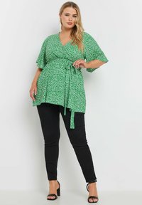 Grön blommig wrap-top med korta fjärilssleeves, V-ringning och knyt i midjan, matchad med svarta smala byxor och klacksandaler.