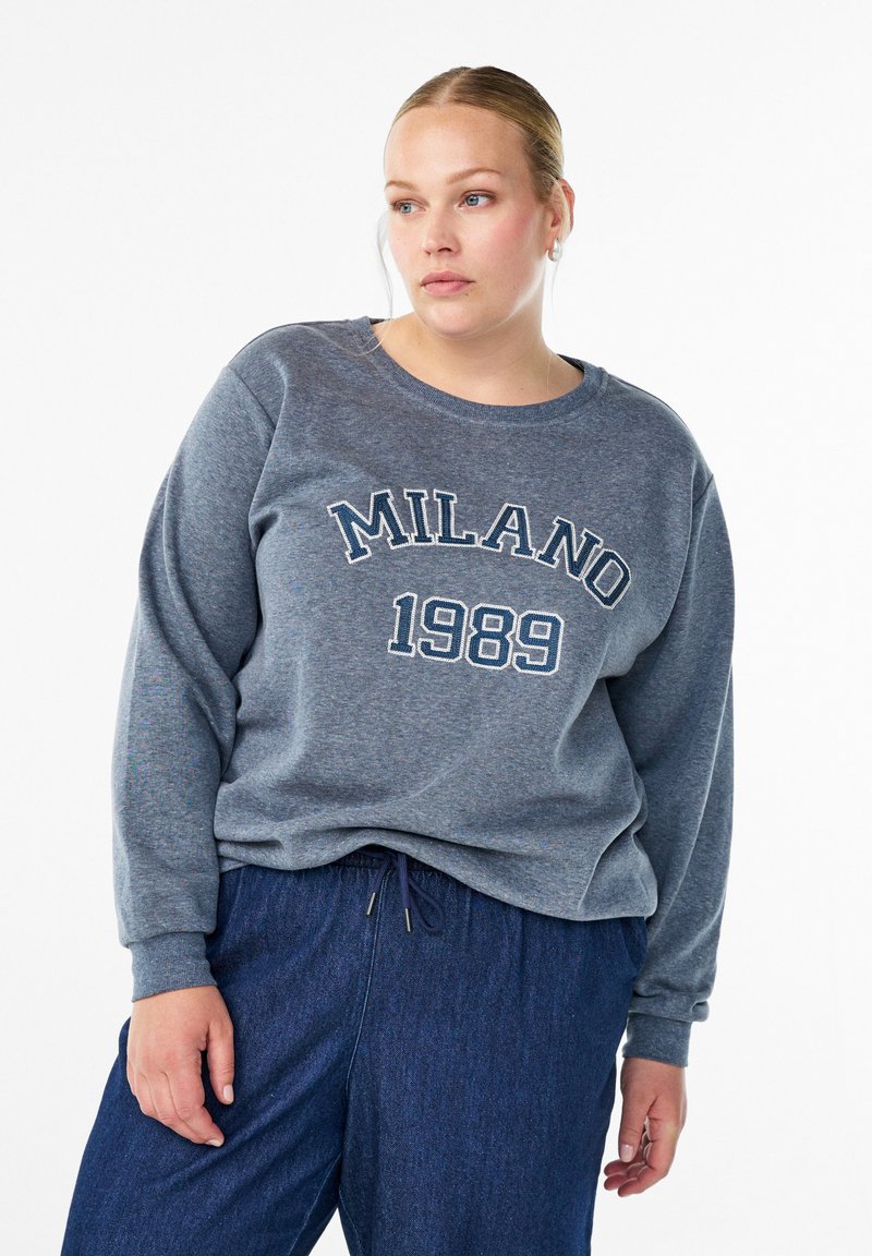 Sivá mikina s textom "MILANO 1989" v svetlom modrom odtieni. Mäkký materiál, okrúhly výstrih a rebrované manžety. Spárovaná s tmavomodrými nohavicami na šnúrku.