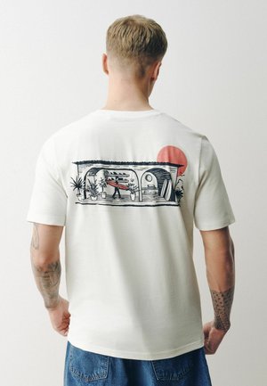 Vyras vilki baltą marškinėlį su grafika, vaizduojančia žmogų nešančią banglentę verandos viduje, apsuptą augalų, o fone matyti raudona saulė.