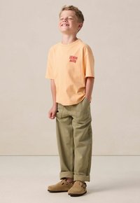 Lichtoranje T-shirt met de tekst "Venice Beach", gecombineerd met losse olijfgroene broeken en tan instappers met een gestructureerd oppervlak.