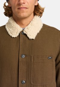 Brown canvas jacket featuring a cream faux shearling collar, two buttons, and a left breast pocket with a black label.