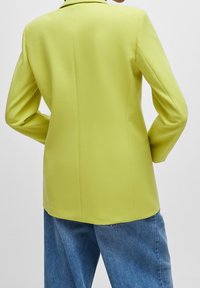 Zieleń limonkowa, dopasowana marynarka o gładkiej teksturze, z wyraźnie zarysowanymi ramionami i subtelnym detalem na tylnym szwie. W połączeniu z luźnymi dżinsami.