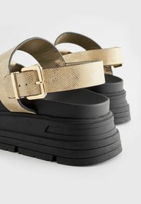 Sandales à plateforme noires avec des brides texturées dorées. Dotées d'une fermeture à boucle, d'un design à semelle superposée et d'une finition en caoutchouc lisse.