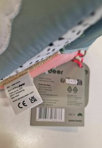 Pehmea plush-mänguasi silt, millel on mitmevärviline tekstuuride segu: roheline kangas, roosa osa ja beež aluspind. Sisaldab hooldusjuhiseid ja ohutuslabelit.