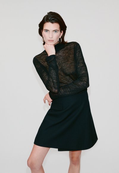 Haut noir transparent à manches longues avec col montant associé à une jupe noire évasée dotée d'une poche latérale, le tout sur fond clair.
