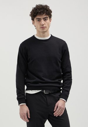 Black knitted sweater with a round neck, ribbed cuffs, and hem. Layered over a white shirt. Model wears black trousers and a belt.