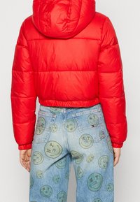 Giacca imbottita rossa con cappuccio, caratterizzata da sezioni trapuntate. Abbinata a jeans azzurri chiari con motivi di faccine sorridenti verdi.