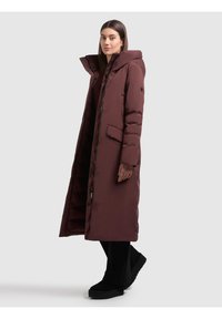 Długa bordowa puchowa kurtka z kapturem, zapinana na suwak i zatrzaski, z bocznymi kieszeniami i pikowaną fakturą. Połączona z czarnymi szerokimi spodniami.