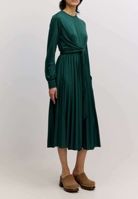 Grünes, gef pleated Kleid mit langen Ärmeln, Bindegürtel und runden Ausschnitt. Kombiniert mit braunen Clogs mit Nieten entlang der Sohle.