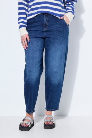 Persona con vaqueros azules, sandalias con hebilla plateada, suéter de rayas azules y blancas, con una mano en el bolsillo y uñas pintadas de morado.