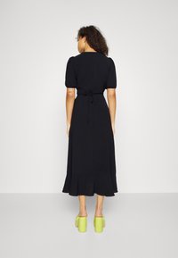 Black, ankle-length dress with a tie waist, puff sleeves, and a flared hem. Worn with bright green chunky heeled sandals.