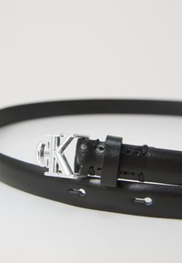 Curea din piele neagră cu cataramă argintie "CK" și multiple găuri de ajustare pe o suprafață albă.