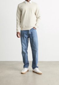 Beige sweatshirt med präglad "BOSS"-logotyp, tillsammans med ljusblå jeans och vita sneakers med bruna sulor. Avslappnad passform och material synliga.