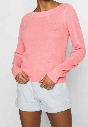 Personne portant un pull en maille rose clair à manches longues et un short bleu clair avec ourlet retroussé, debout les mains près de la taille.