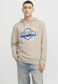 Lysbeige hoodie med frontlogo-design, snoretræk i hætten og ribbede manchetter. Båret med lyseblå denim jeans. Enkel, afslappet stil.