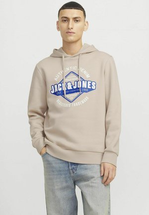 Light beige hoodie with front logo design, drawstring hood, and ribbed cuffs. Worn with light blue denim jeans. Simple, casual style.