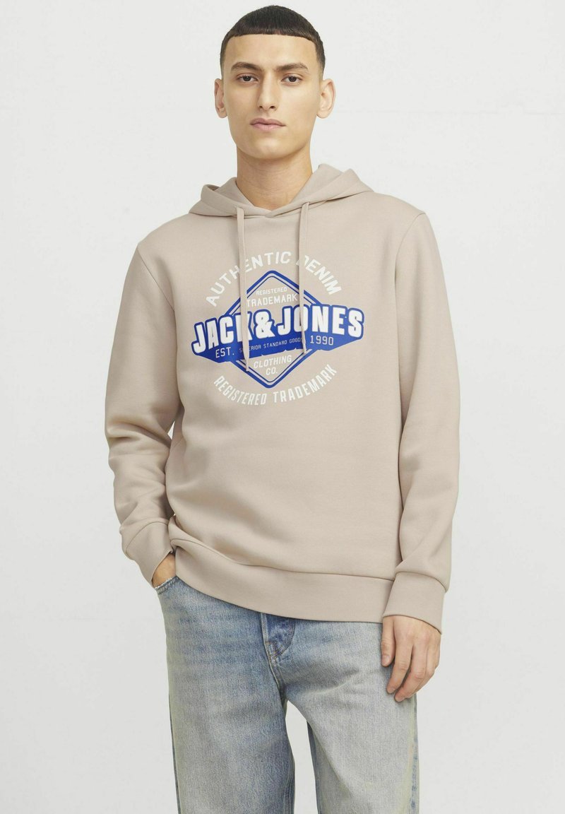 Lichtbeige hoodie met logo-ontwerp aan de voorkant, trekkoordkap en ribgebreide manchetten. Draagt met lichtblauwe denim jeans. Eenvoudige, casual stijl.