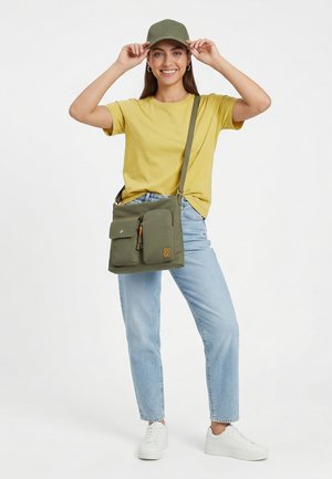 Junge Frau lächelnd, trägt ein gelbes T-Shirt, eine olivgrüne Kappe, hellblaue Jeans, weiße Sneakers und trägt eine olivgrüne Umhängetasche.