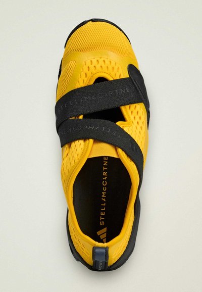 Zapato deportivo amarillo con parte superior de malla transpirable, correas negras ajustables, suela texturizada y detalles del logo en el interior.