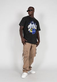 Camiseta gráfica negra con estampado colorido, combinada con pantalones cargo beige con múltiples bolsillos y zapatillas blancas. Las gafas completan el look.