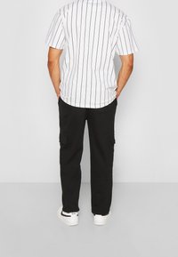 Mężczyzna w czarnych spodniach cargo, białych sneakersach Nike i białej koszuli z krótkim rękawem w czarne pionowe paski, stojący z rękami w kieszeniach.