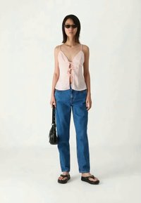 Sieviete, kas valkā saulesbrilles, gaiši rozā bez piedurknēm topu ar priekšu saistītām saitēm, zila džinsu bikses, melnas sandales un tur rokās melnu somu.