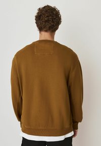 Sweatshirt din bumbac maro, cu o patch texturat pe spate, manșete și tiv cu nervuri. Are un guler tip crew și o croială lejeră.