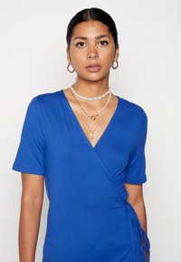 Vestido azul de manga curta em estilo envolvente com decote em V, apresentando detalhes pregueados e laços de lado. Textura de tecido suave.