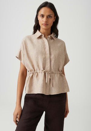 Vrouw draagt een beige kortemouw-blouse met knopen en een aantrekkoord in de taille en een donkere broek, staand tegen een effen lichte achtergrond.