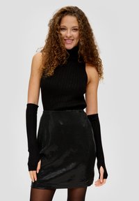 Schwarzes, geripptes Rollkragenshirt, kombiniert mit einem glänzenden, schwarzen Pailletten-Minikleid. Lange fingerlose Handschuhe reichen von den Ärmeln des Shirts.