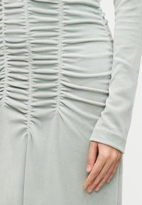 Hellgrünes, langärmliges Kleid mit Raffung entlang des Körpers und der Ärmel, aus weichem, strukturiertem Stoff. Glatte, figurbetonte Silhouette.