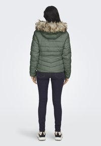 Grønn polstret jakke med en avtakbar hette i falsk pels, med kviltet tekstur og en svakt innsvingt silhuett. Båret med mørke jeans.