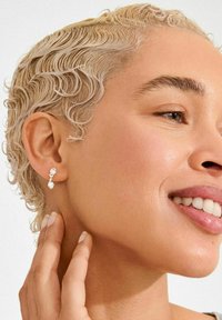 Orecchini a bottone in tono argento con due pietre preziose trasparenti a forma di goccia e tonda, su uno sfondo chiaro.