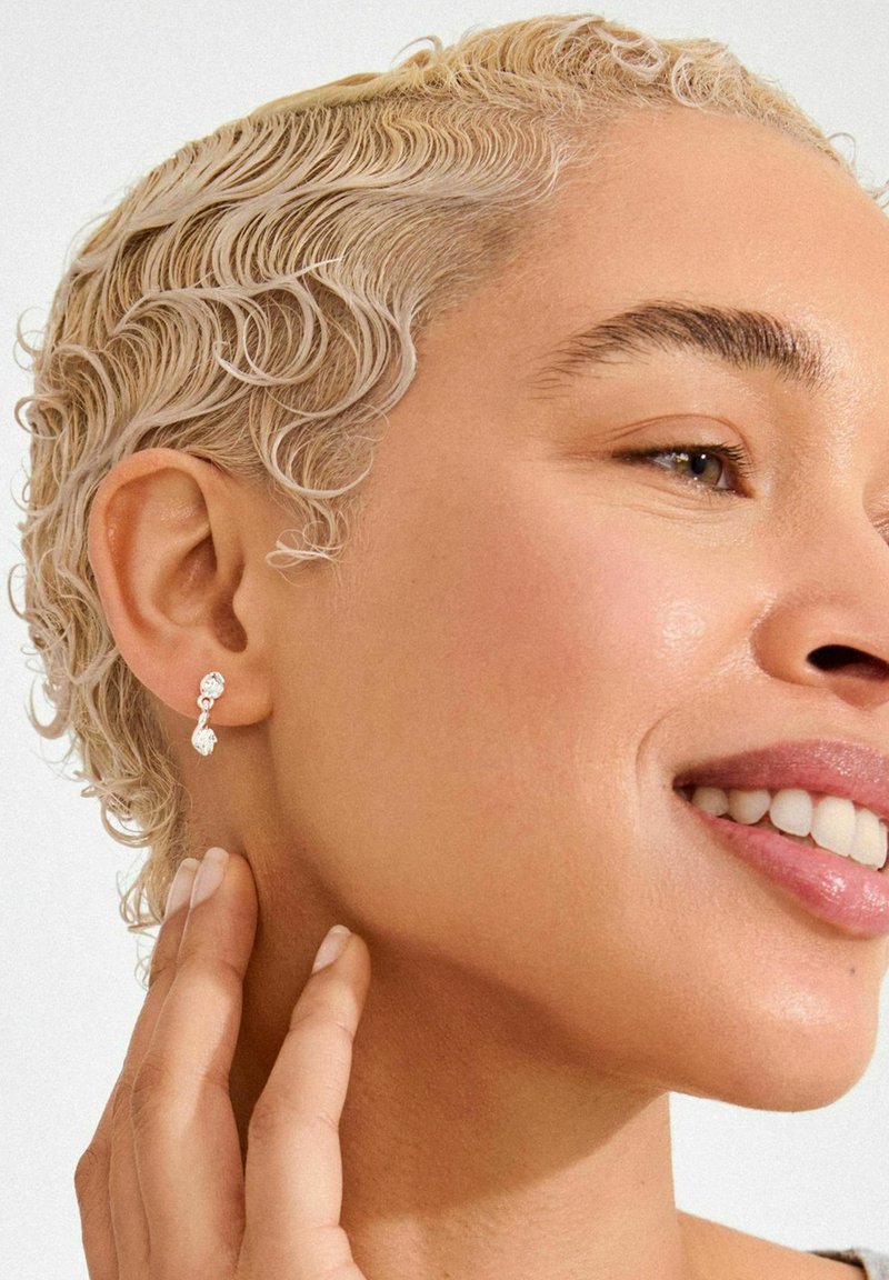 Orecchini a bottone in tono argento con due pietre preziose trasparenti a forma di goccia e tonda, su uno sfondo chiaro.