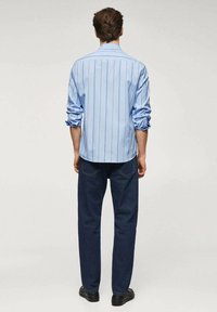 Camisa a rayas azul claro con mangas enrolladas, combinada con jeans azul oscuro y zapatos negros. La vista trasera muestra los detalles y el ajuste de la camisa.