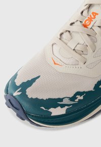 Hardloopschoen met een beige mesh bovenwerk, teal accenten en een gestructureerde zool. Opvallend oranje logo en abstract patroon op de neus.