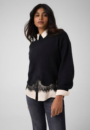 Femme aux longs cheveux foncés portant un pull noir avec un ourlet en dentelle par-dessus une chemise blanche et un jean foncé, debout devant un fond uni.