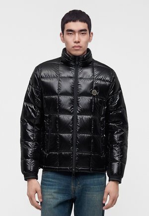 Junger Mann trägt eine glänzende schwarze gesteppte Daunenjacke mit hohem Kragen, Reißverschluss vorne und blaue Jeans vor einfachem hellem Hintergrund.
