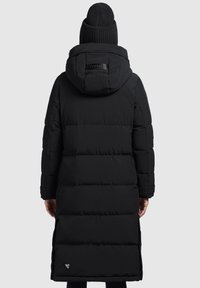 Zwarte pufferjas met capuchon, lange lengte, horizontaal gewatteerd patroon, zijsplitten en een merklabel op de rug nabij de nek. Gebreide zwarte beanie.