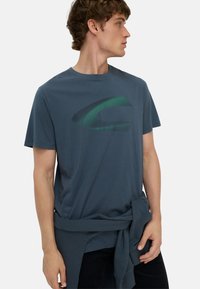 Mørkeblå bomulds t-shirt med korte ærmer og et grønt grafisk logo på forsiden. Har en afslappet pasform og en knyttet stofdetalje i taljen.