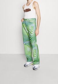 Barevné kalhoty s motivem zelené a modré barvy, volného střihu, spárované s bílým tílkem. Kalhoty mají vlnité pruhy a jsou vyrobeny z látky.