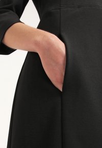 Tissu de robe noire à texture lisse avec une poche latérale. Une main est placée dans la poche, mettant en valeur le design pratique de la robe.