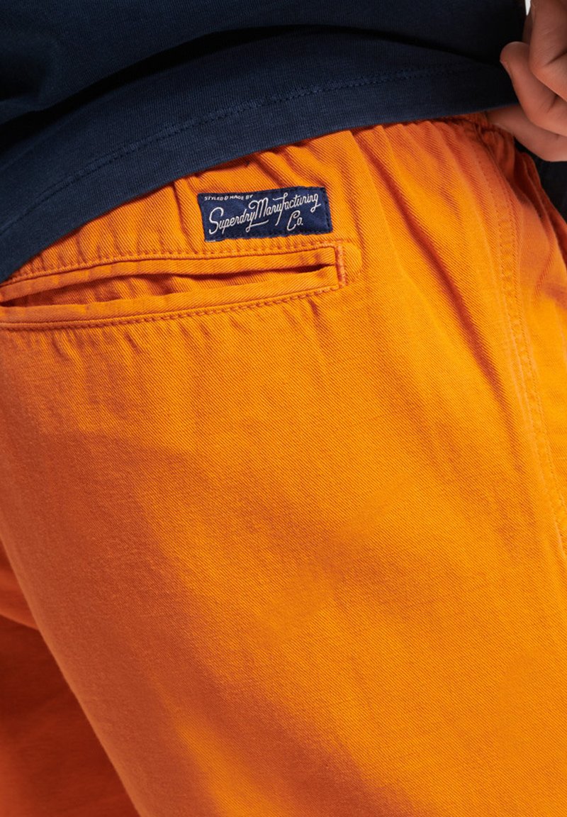 Pomarańczowe bawełniane spodnie o gładkiej fakturze, z tylną kieszenią i niebieską metką z białym napisem "Superdry Manufacturing Co."
