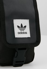 Borsa di stoffa nera con un logo Adidas bianco applicato, fissata da una fibbia nera. Presenta una forma rettangolare e una superficie texture.