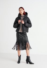 Doudoune noire, tissu texturé, design avec capuche ; pull gris en dessous. Jupe en cuir avec des franges verticales ; bottines noires.