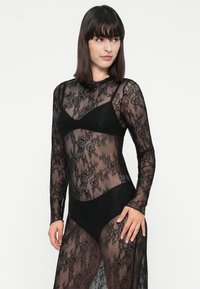 Žena v černých květinových krajkových šatech s dlouhým rukávem a odpovídajícími černými spodními prádly, která se mírně dívá na stranu proti jednoduchému pozadí.