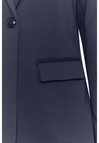 Blazer bleu marine avec une poche à rabat et un bouton noir visible sur le côté gauche devant.