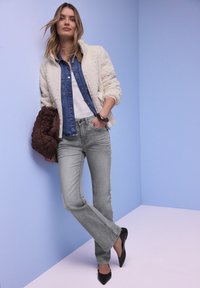Jeune femme en jean gris clair, chemise en denim bleu, veste en polaire blanche, tenant une veste matelassée marron, debout contre un mur bleu pâle.